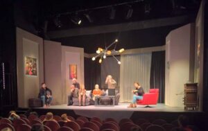 Na scenie teatralnej piątka aktorów siedzi na kanapie. Po prawej, na fotelu, tłumaczka języka migowego.