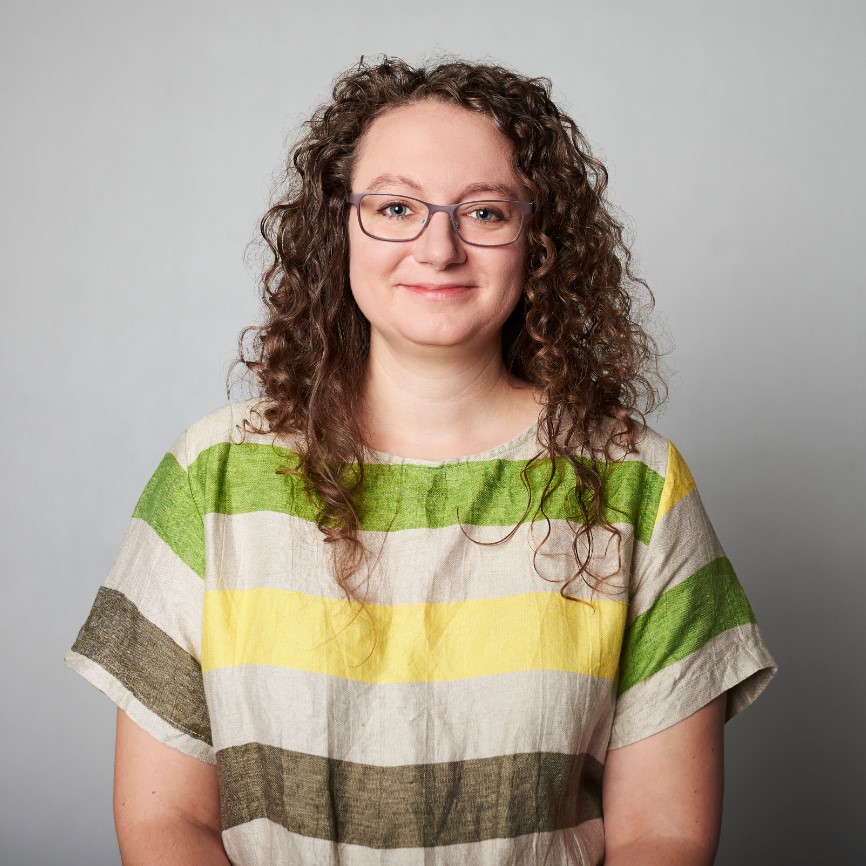 Zdjęcie portretowe: Magda Brumirska-Zielińska. Długie, kręcone włosy, na nosie okulary. Ubrana w koszulkę w kolorowe paski. Patrzy w obiektyw i uśmiecha się.