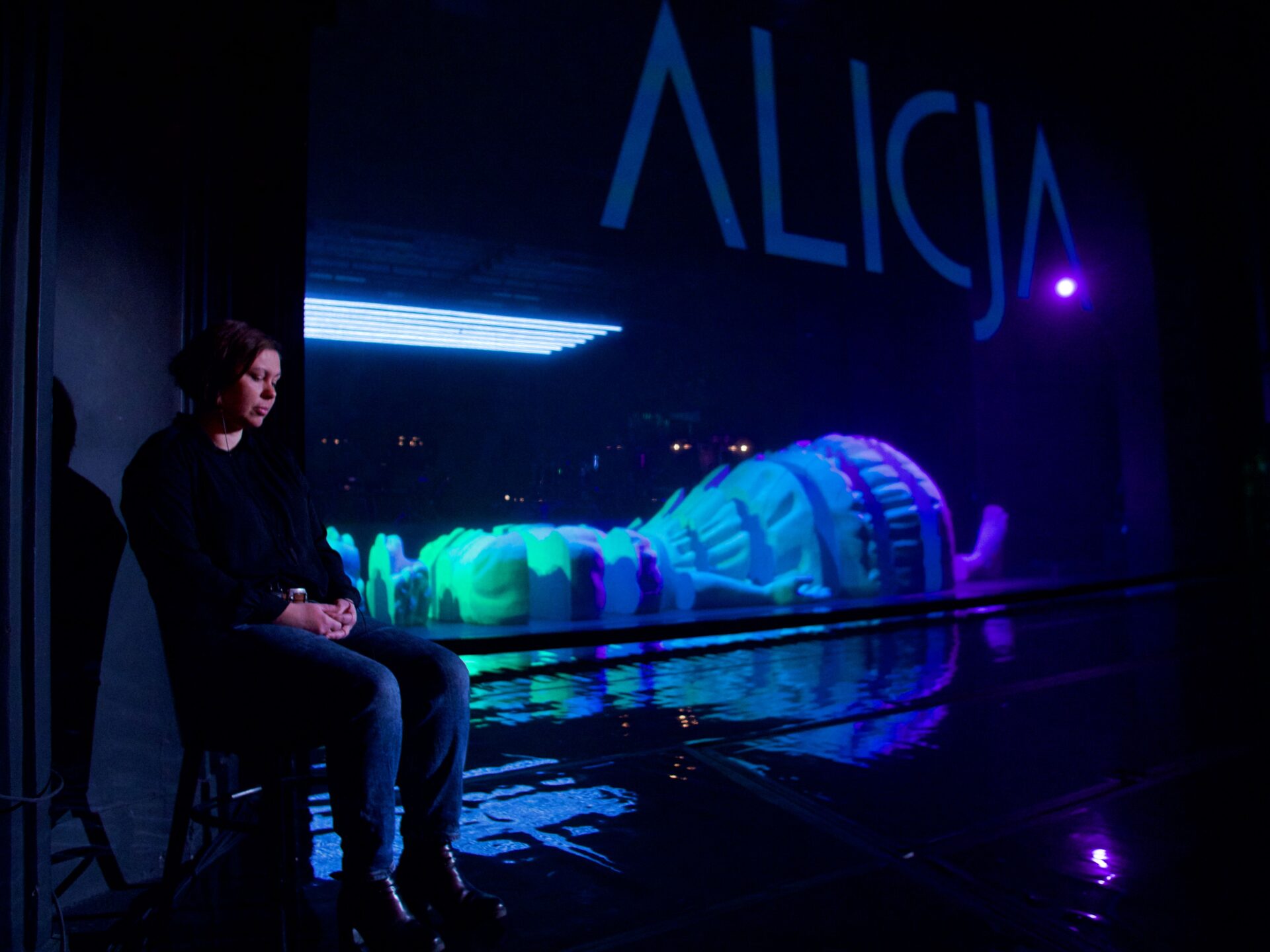 Scena teatralna. Tłumaczka Ela Resler siedzi na krześle. W tle dekoracje i wielki napis Alicja.